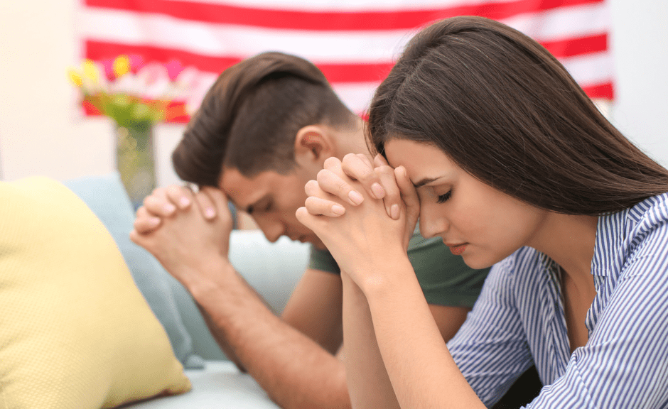 An image of a young white couple praying in front of a US flag to introduce an article about how conservative Christians downplay Christian nationalism
